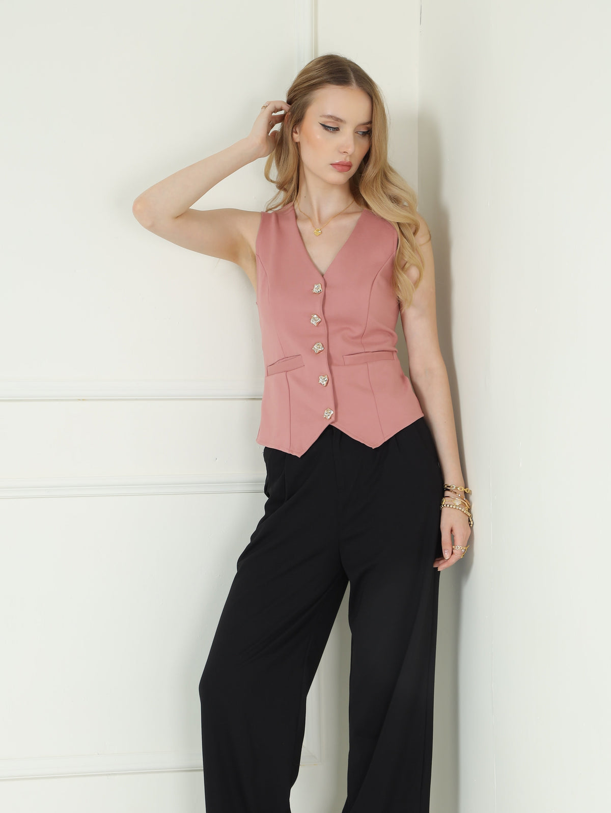 A woman models a sleeveless dusty pink vest with gold buttons and black wide-leg trousers, standing in a white paneled room corner.