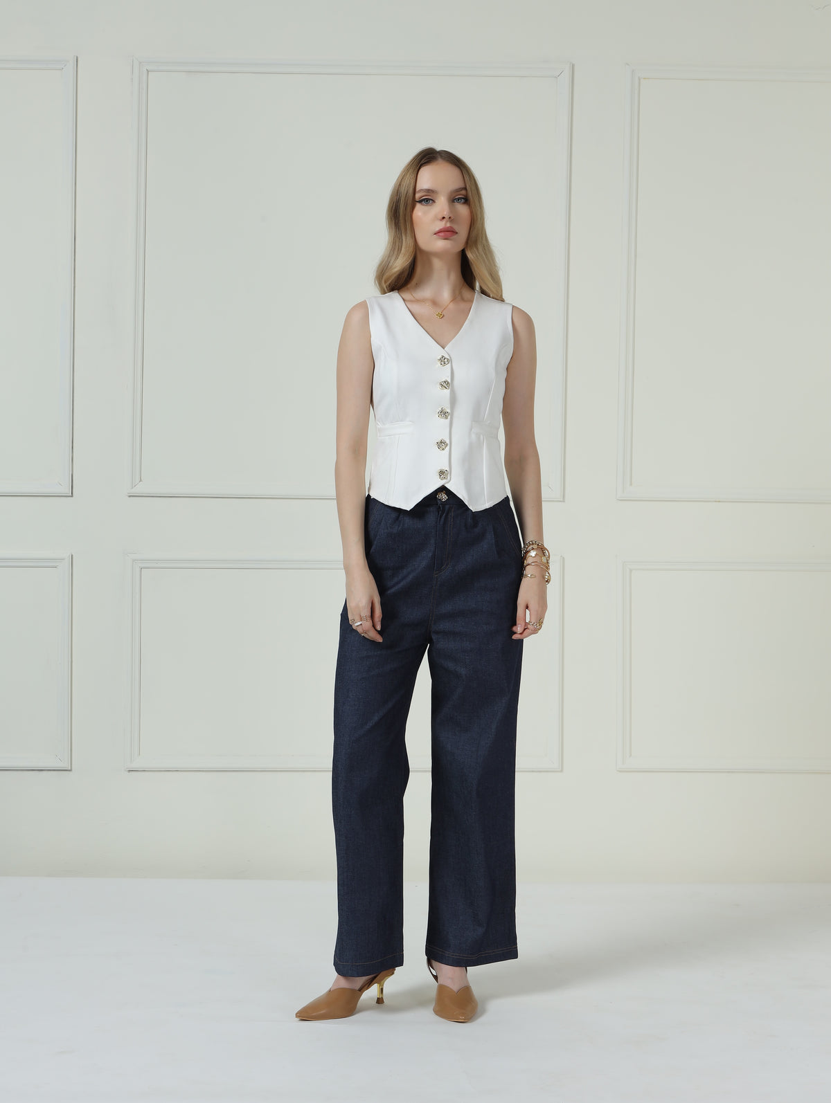 A woman stands in a studio wearing a sleeveless white vest with gold buttons, dark navy wide-leg trousers, and tan pointed-toe heels against a cream paneled wall.