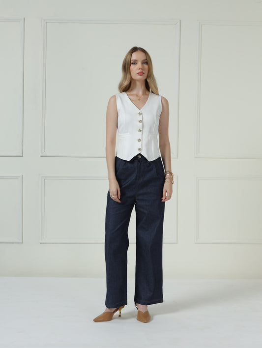 A woman stands in a studio wearing a sleeveless white vest with gold buttons, dark navy wide-leg trousers, and tan pointed-toe heels against a cream paneled wall.