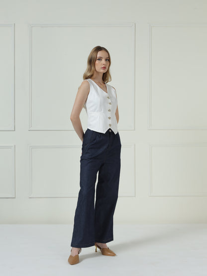 A woman models a sleeveless white vest with gold buttons and dark navy wide-leg trousers, standing against a white paneled wall.